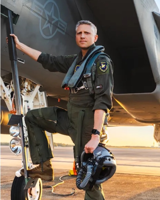 Tucker Hamilton in a flight suit beside a fighter jet at sunrise, helmet in hand.